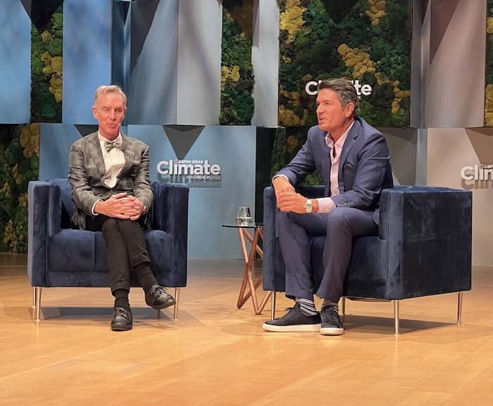 Bill Nye (left) and Louis Aguirre (right) sit on blue chairs on a wood stage. 