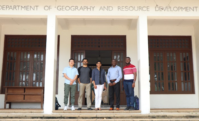 Five people standing between two white columns in front of three dark wood window and door frames. Above the people, a sign reads Department of Geography and Resource Development.